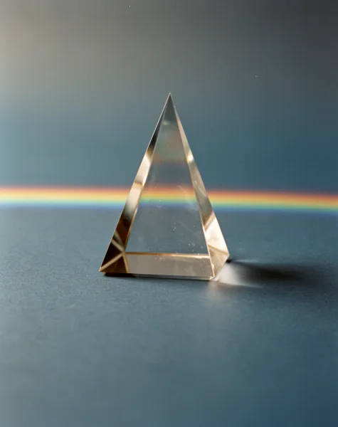 A clear glass prism on a dark surface with a rainbow reflection in the background, under soft lighting.