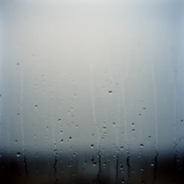 Raindrops streaming down a foggy window pane with a blurred background in soft blue and gray tones.