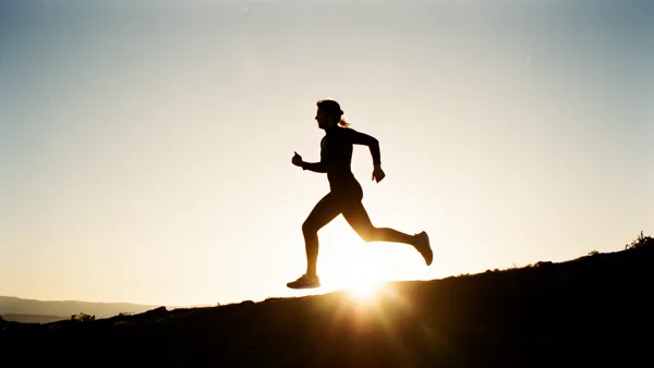 Silhouette of a person running up a hill at sunset with the sun low on the horizon, casting a warm glow.