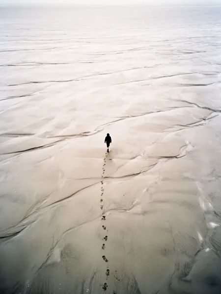 Aerial view of a person walking on a vast sandy beach with footprints trailing behind under an overcast sky.
