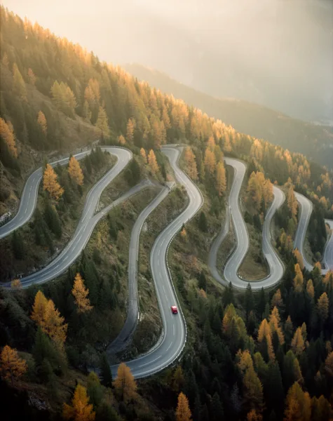 Aerial view of a winding mountain road with a red car, surrounded by autumnal trees in soft, diffused light.