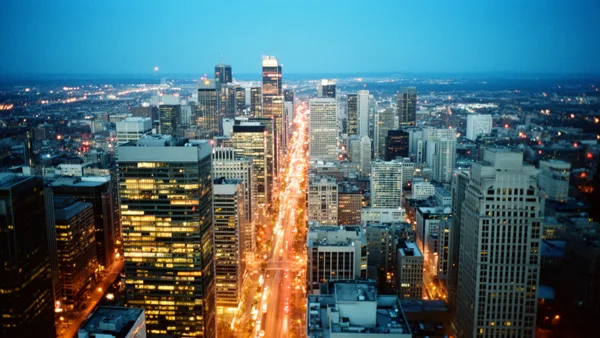 Aerial view of a city skyline at dusk with skyscrapers and a brightly lit avenue under a blue twilight sky.