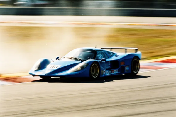 A blue race car with the number 63 on a track, captured in motion with a blurred background and bright natural lighting.