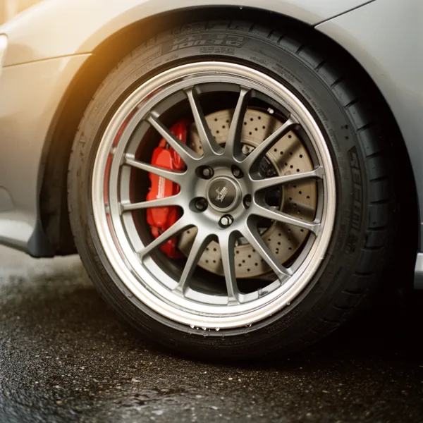 Close-up of a sports car wheel with alloy rim and red brake caliper on a wet road surface in warm lighting.