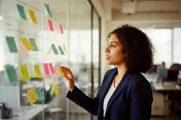 A businesswoman in a blue blazer arranging colorful sticky notes on a glass wall in a modern office with natural light.