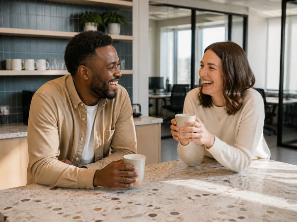 Two colleagues laughing and holding coffee mugs at a stone countertop in a modern office with sunlight streaming in.