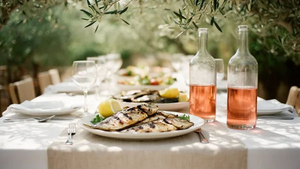 Outdoor dining table with grilled fish, lemons, and bottles of rosé wine under olive trees with soft natural light.