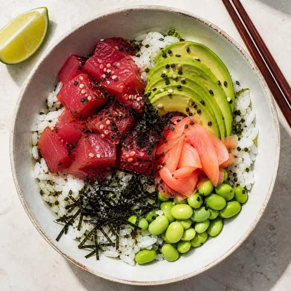 A tuna poke bowl with tuna sashimi, avocado, edamame, pickled ginger, and nori on rice, garnished with sesame seeds, under natural light.