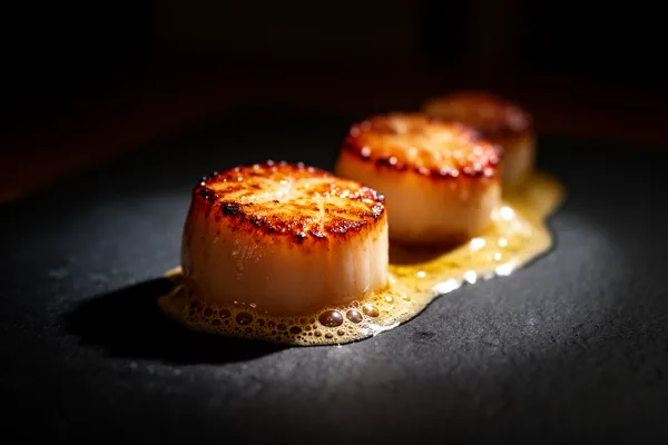 Three seared scallops with golden tops and buttery sauce on a dark slate plate under dramatic lighting.