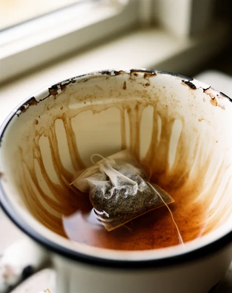 A close-up of a stained teacup with a teabag inside, showing tea residue and a chipped rim, lit by natural window light.