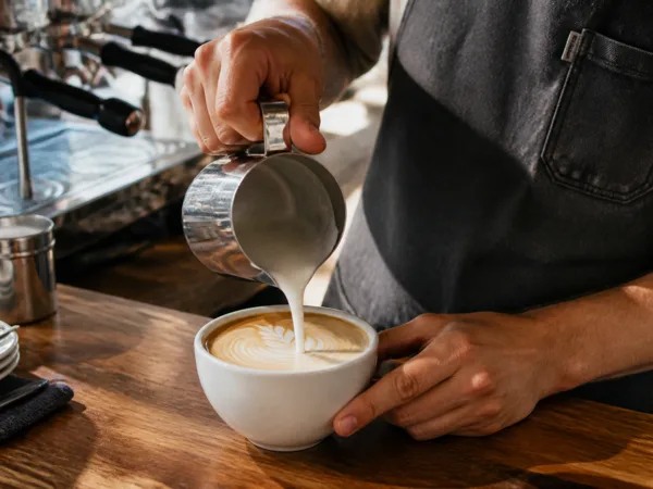 A barista pours steamed milk into a cup of espresso, creating latte art on a wooden counter with an espresso machine in the background.
