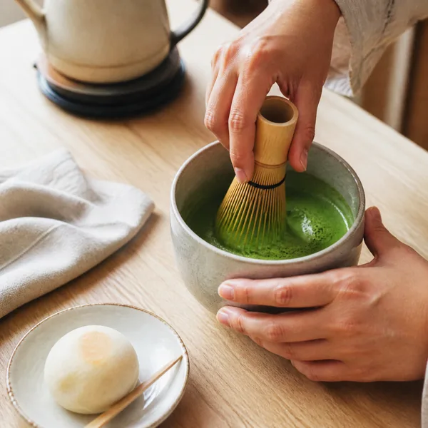 Hands using a bamboo whisk to prepare matcha tea in a ceramic bowl on a wooden table with a teapot and a Japanese confection nearby.
