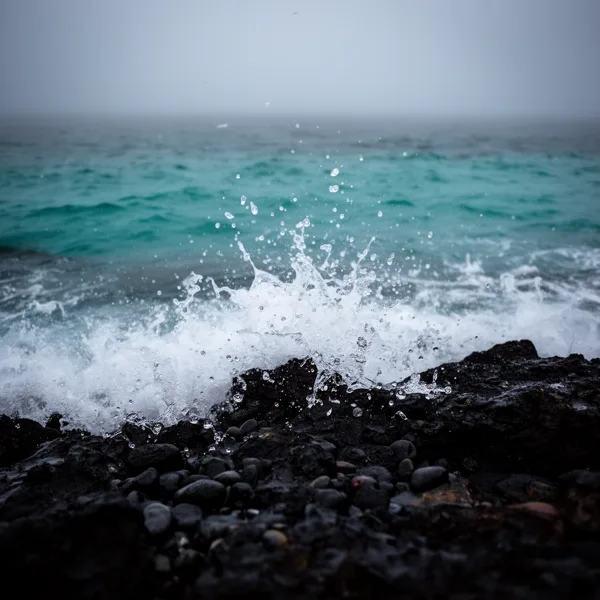 Waves crashing on dark rocky shore with turquoise sea and overcast sky, water droplets suspended in air.