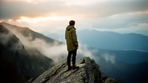 A hiker in a green jacket stands on a rocky mountain peak, looking at distant misty mountains under a glowing sunset sky.