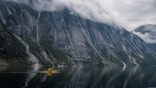 A kayaker in a yellow jacket paddles on a calm lake with towering cliffs and misty clouds in the background under soft light.