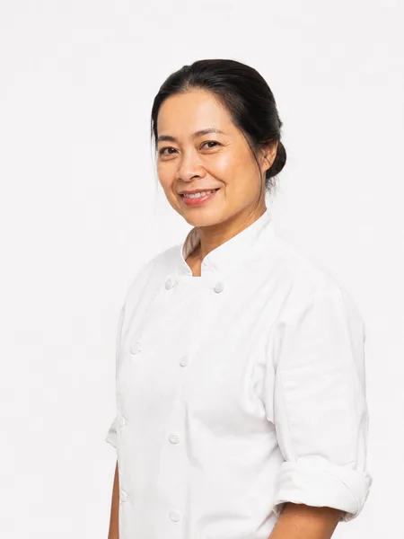 A female chef in a white uniform smiling against a white background with her hair tied back.