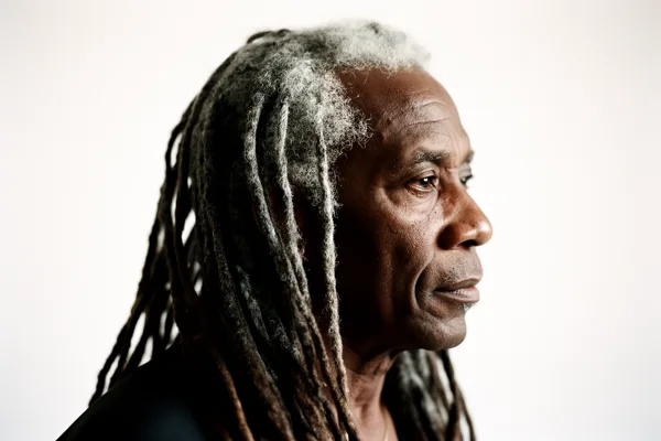 Profile of an elderly man with long gray dreadlocks against a white background, lit with soft natural light.