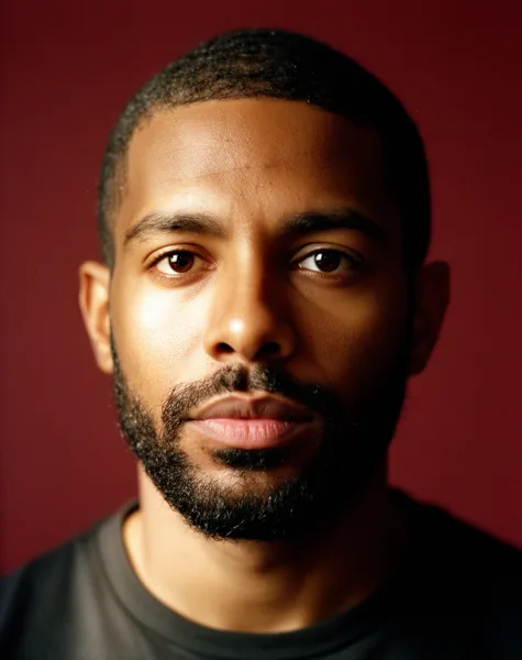 Close-up portrait of a man with a beard and short hair against a burgundy background with soft lighting.