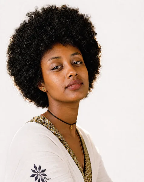 Portrait of a woman with afro hair wearing an embroidered white garment and a choker necklace, set against a neutral background with soft lighting.
