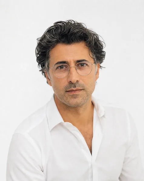 A mature man with glasses and salt-and-pepper hair wearing a white shirt, against a white background with soft lighting.