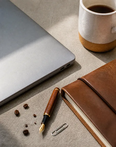 A laptop, coffee mug, leather notebook, fountain pen, and coffee beans on a fabric surface with soft natural light.