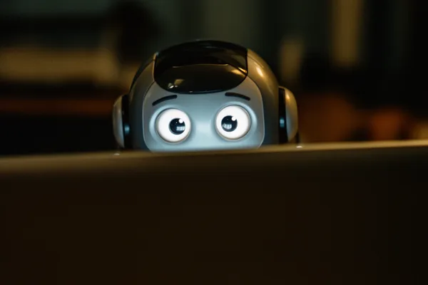 A small robot with glowing eyes peeking over a screen in a dimly lit room.