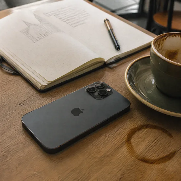 A smartphone, open notebook with a pen, and a coffee cup on a wooden table with soft natural lighting.