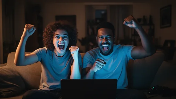 Two excited people sitting on a couch, cheering while looking at a laptop screen in a dimly lit room.
