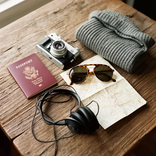 Travel essentials including a camera, passport, sunglasses, map, sweater, and headphones on a rustic wooden table with natural light.