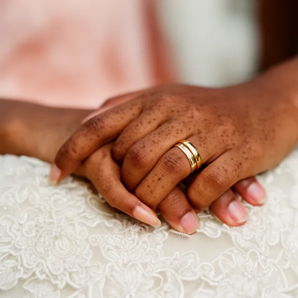 Close-up of a bride's hands with a gold wedding ring resting on a lace wedding dress in soft natural light.