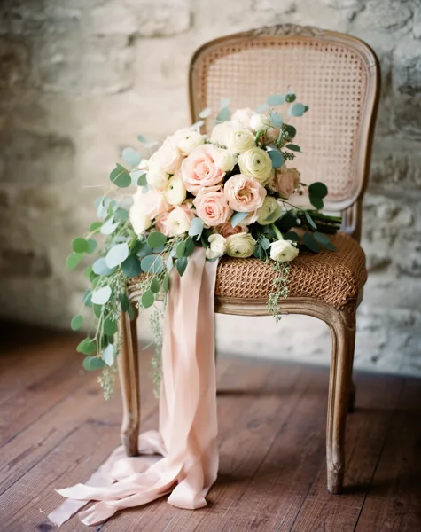 A wedding bouquet with pink and cream roses on a vintage wicker chair against a stone wall, with soft natural lighting.